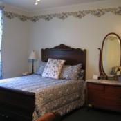 guest room; white walls with floral banner around ceiling, small wooden bedframe, floral bedspread, wooden dresser with mirror
